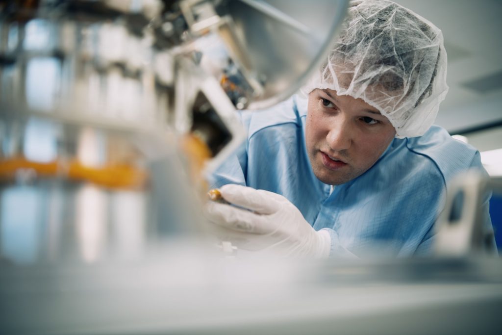 Engineer in a cleanroom environment wearing a hairnet, gloves, and protective blue gown, carefully inspecting or assembling high-precision equipment.